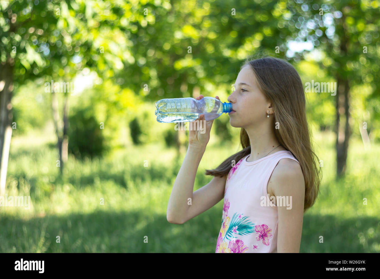 Una giovane ragazza bionda in una t-shirt rosa beve l'acqua da una bottiglia di plastica su uno sfondo di colore verde foresta. Ragazza giovane beve l'acqua Foto Stock