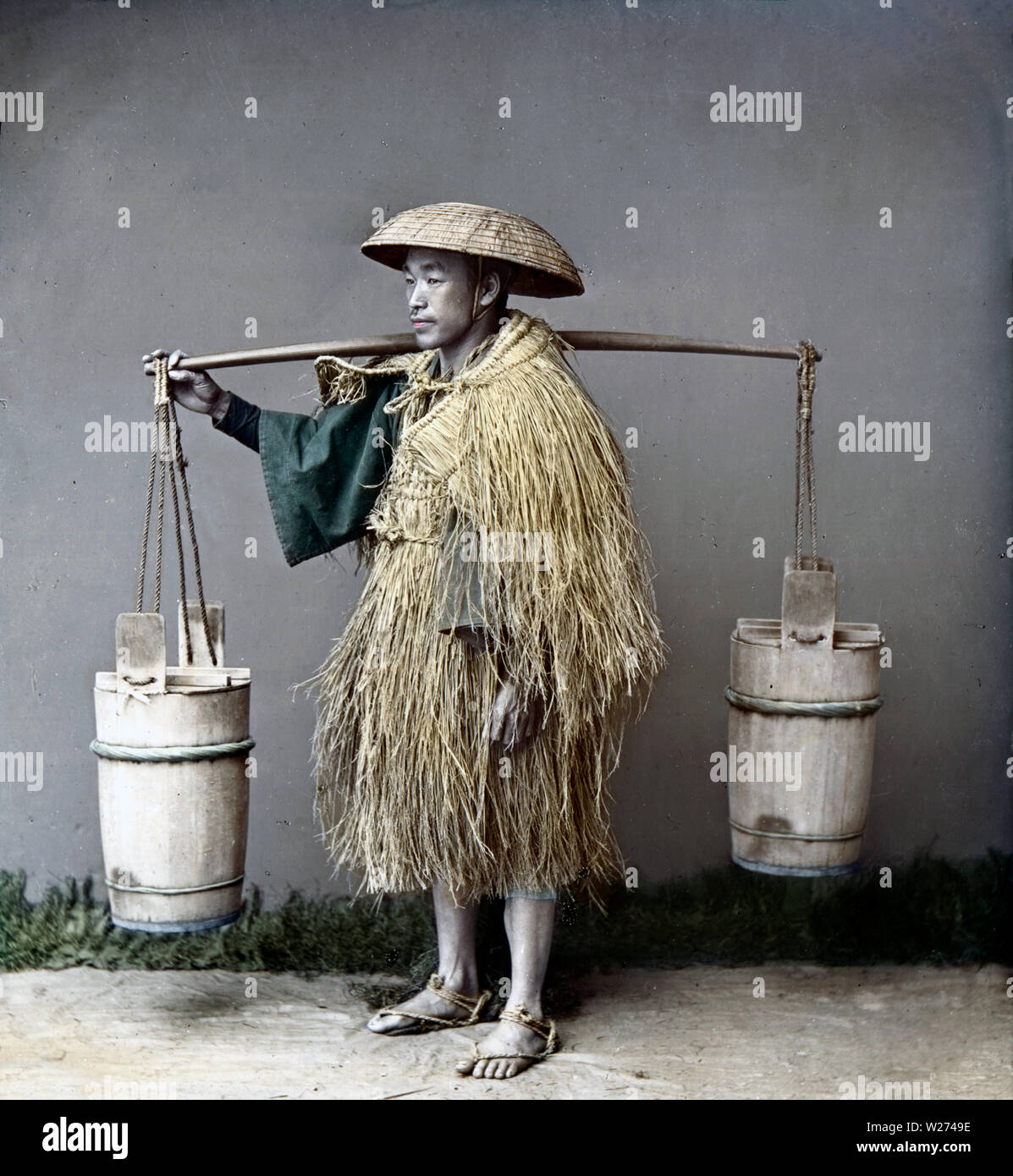 1880 Giappone - Contadino con benne ] - Un agricoltore giapponese indossa  un mino (paglia impermeabile) e un sugegasa cappello conico sta portando un  palo con benne di (probabilmente) notte suolo.