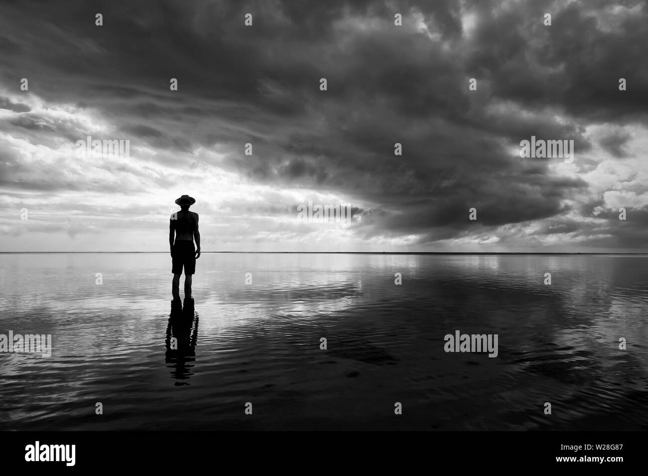 Moody monocromatica di silhouette uomo in hat in piedi di calmare le acque poco profonde riflettente skyscape drammatico sul mare orizzonte Foto Stock