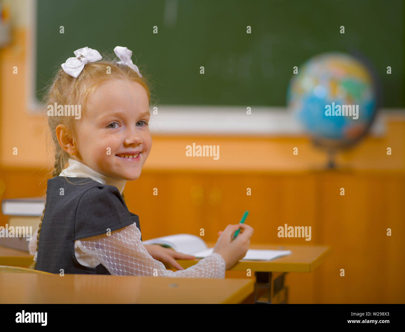 La scolaretta sorridente si siede a una scrivania in aula sullo sfondo del bordo della scuola Foto Stock