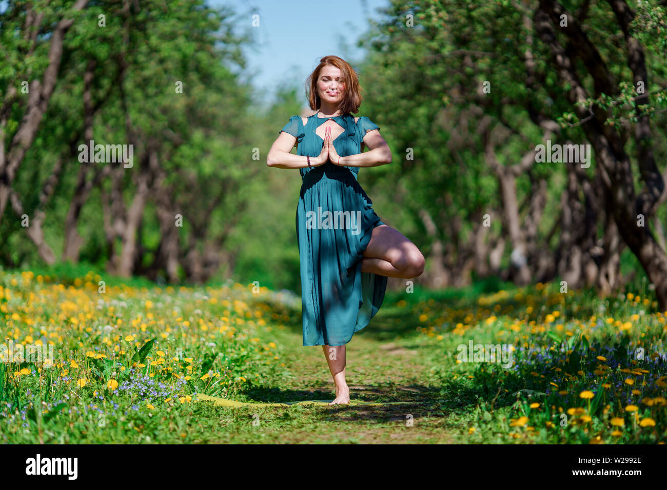 Foto della ragazza con gli occhi chiusi in lungo abito verde facendo yoga in foresta Foto Stock