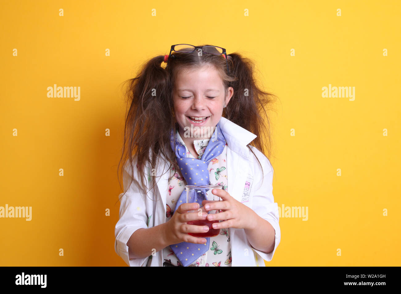 Signor - Scienza Geek giovane ragazza / femmina - 7 anni - bambino ridere per fotocamera, gli occhi chiusi, tenendo in borosilicato scienza becher su sfondo giallo Foto Stock