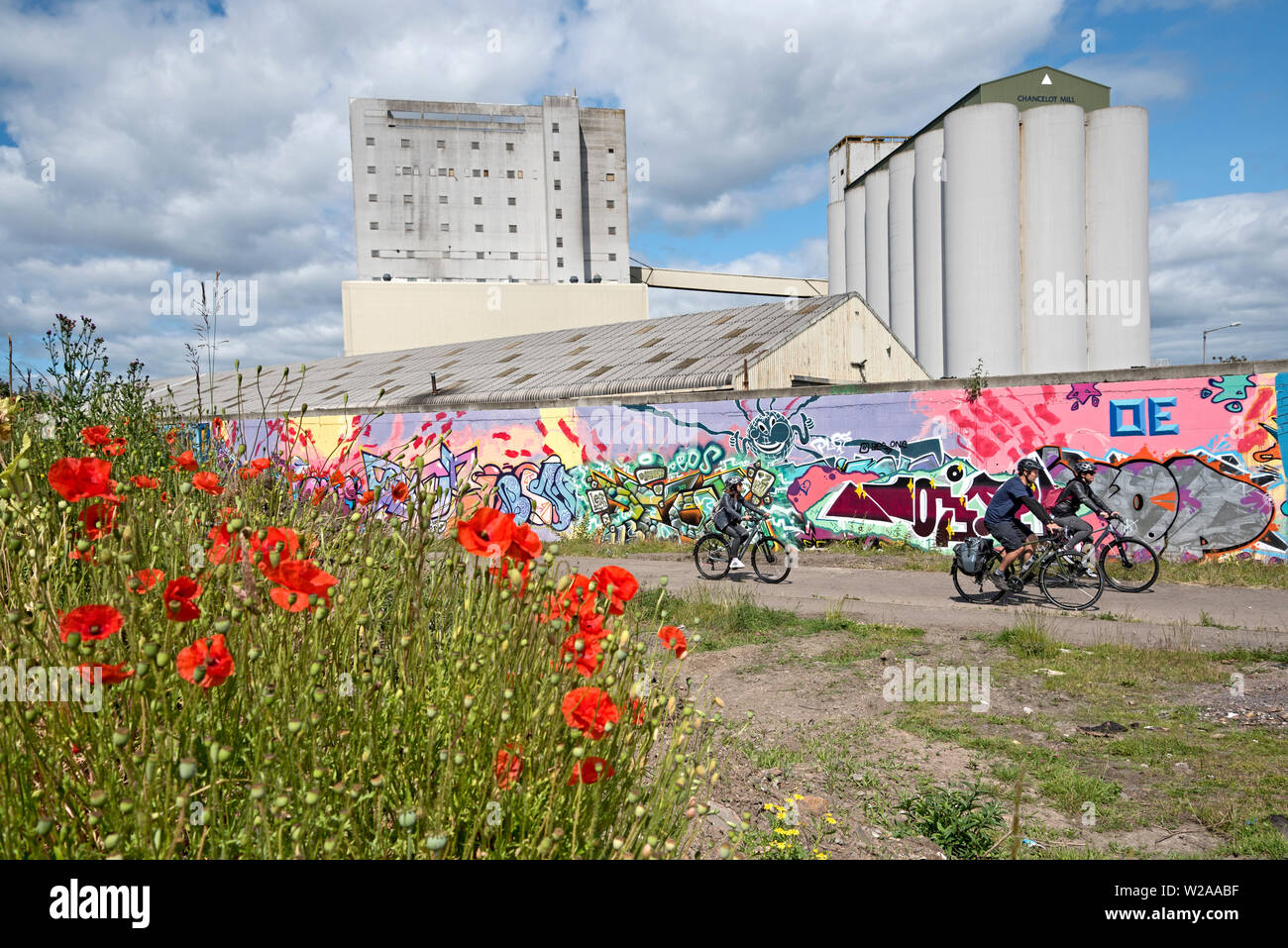 Un gruppo di ciclisti passando graffiti, papaveri selvatici e Chancelot mulino di farina in Leith, Edimburgo, Scozia, Regno Unito. Foto Stock