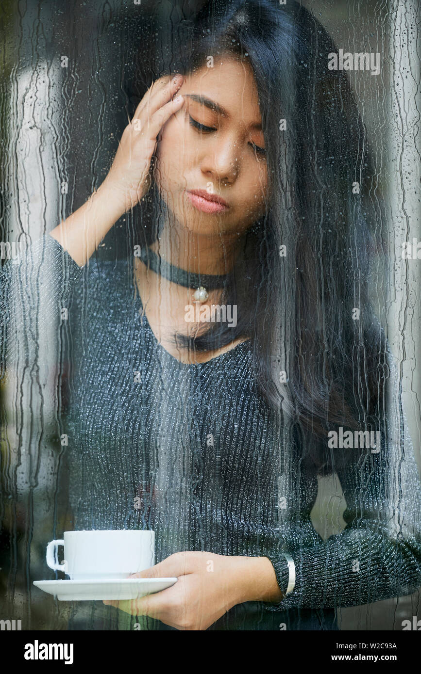 Triste Asian giovane donna in piedi vicino alla finestra e bicchiere di contenimento di caffè piove dietro la finestra e lei sembra turbare Foto Stock