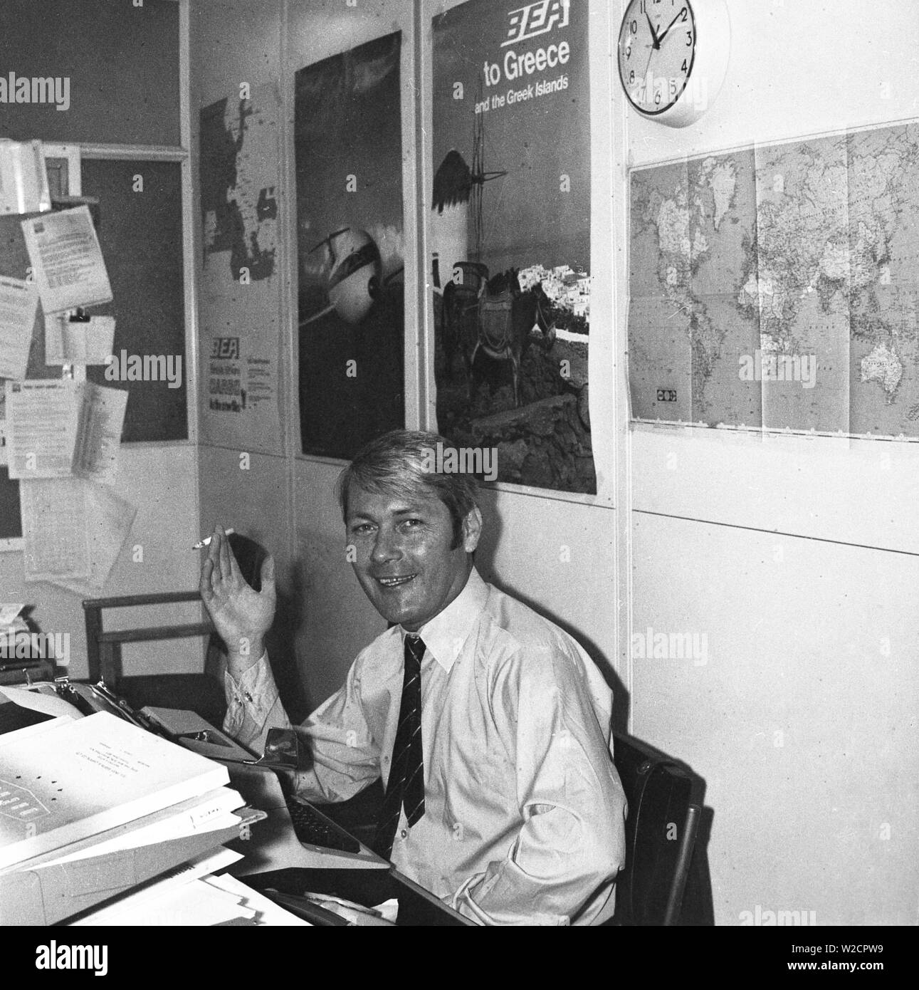 1974, storico, un uomo che lavora in ufficio di un agente di viaggio, seduto alla sua scrivania, godendo di una sigaretta, Inghilterra, Regno Unito. Foto Stock