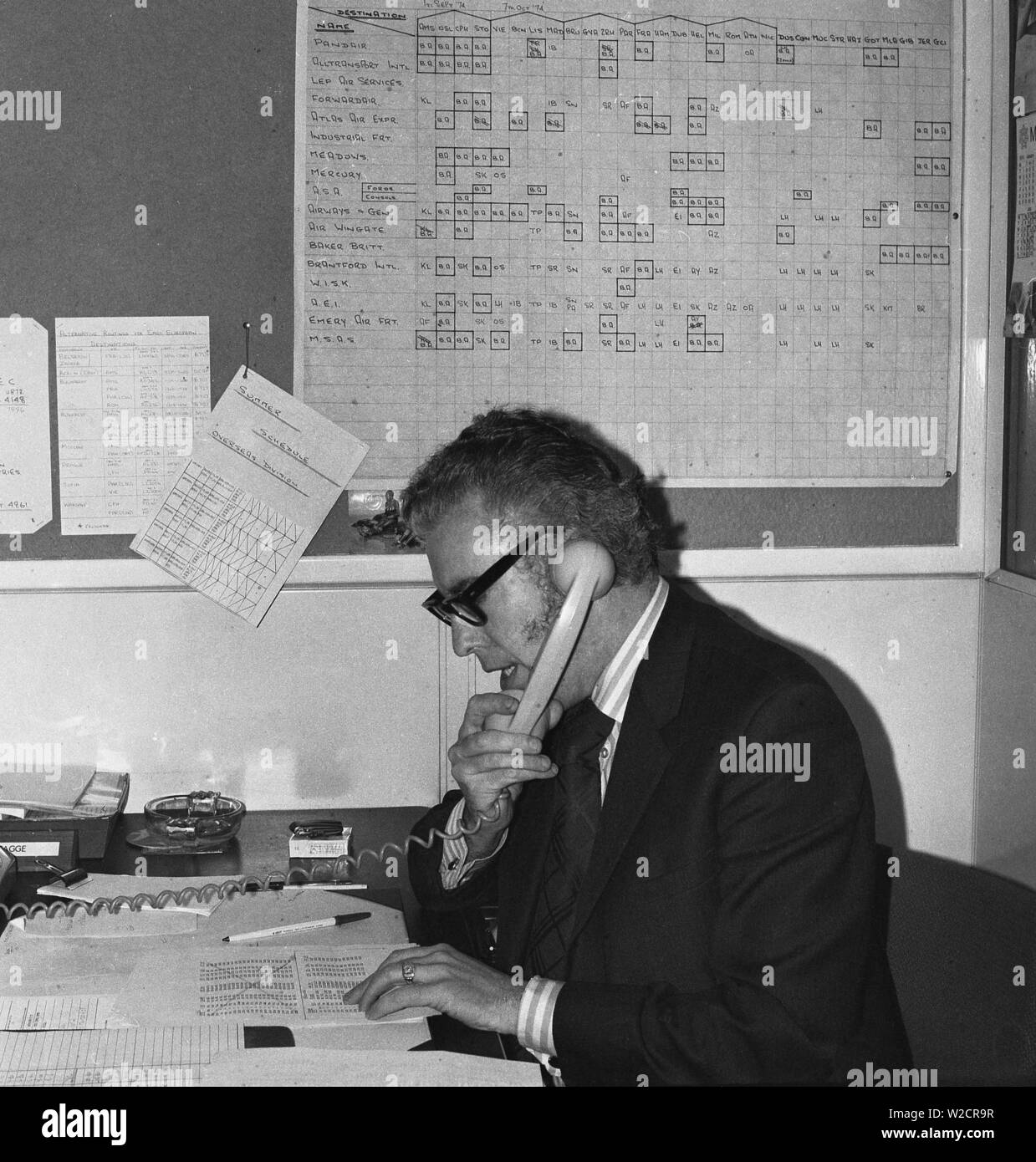 1974, storico, un uomo in una tuta lavora in ufficio di un freight forwarding agency, parlando al telefono, England, Regno Unito, con un posacenere e accendisigari e un pacchetto di sigarette besiide home sulla scrivania. Foto Stock