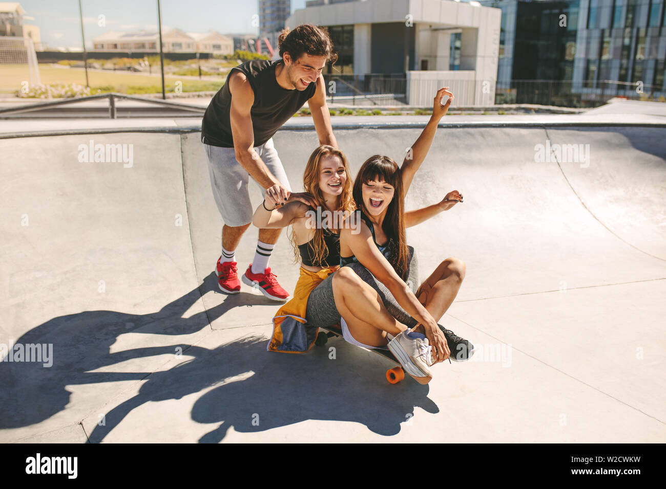 Felice giovane spingendo due ragazze seduta su skateboard a skate park. Gruppo di amici di godere in una ciotola di pattino. Foto Stock