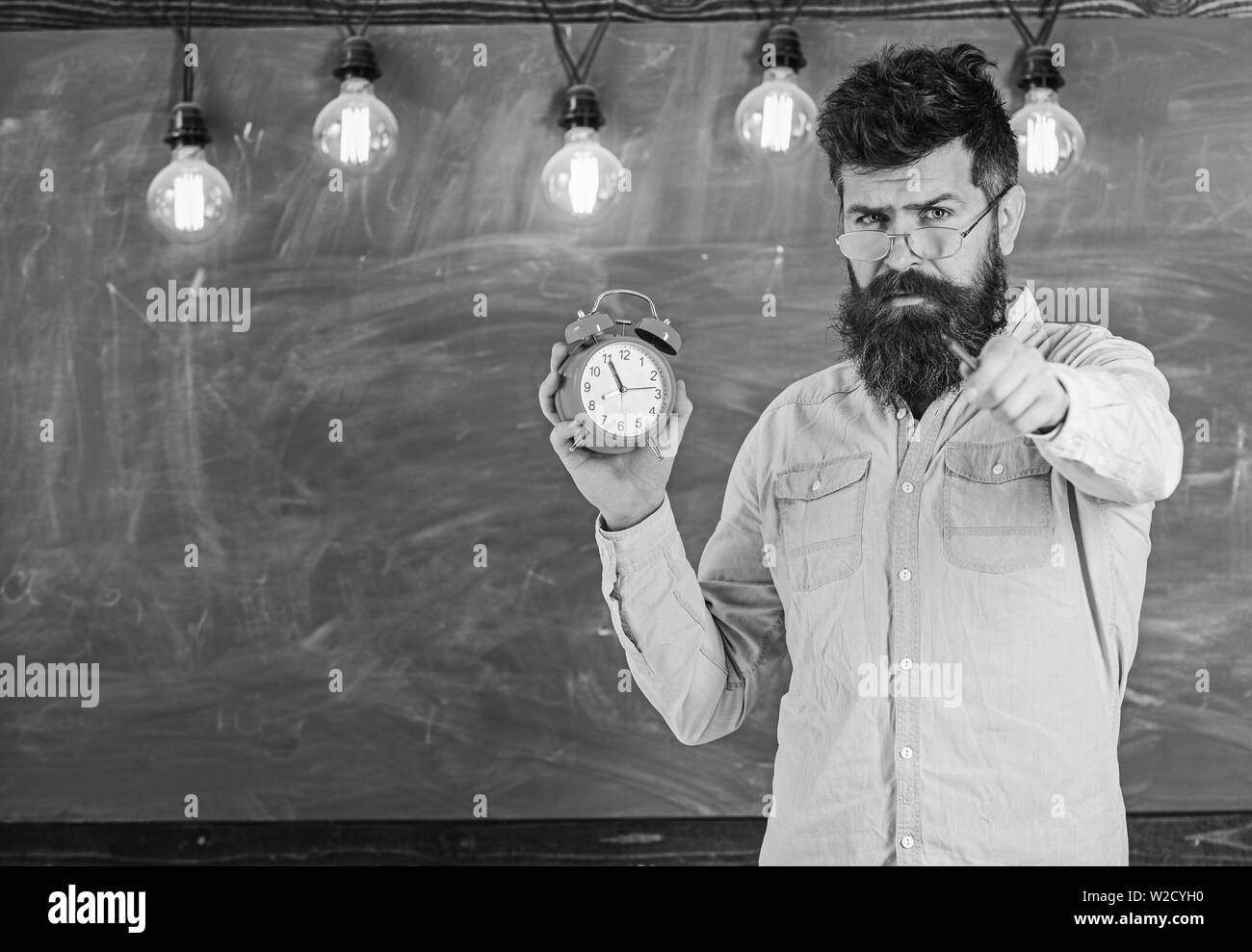 Barbuto hipster detiene orologio, lavagna su sfondo, copia dello spazio. Uomo con barba e baffi sul rigoroso volto. Insegnante in occhiali tiene sveglia e rivolta in avanti. Concetto di disciplina. Foto Stock