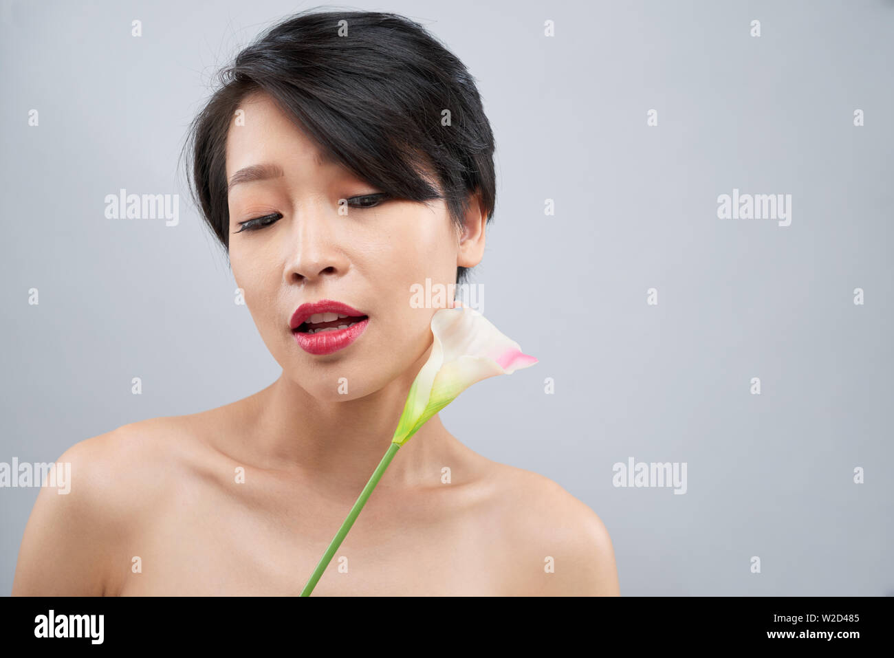 Ritratto di Asian ragazza giovane con corti capelli scuri e con belle make-up in posa con fiore contro lo sfondo grigio Foto Stock