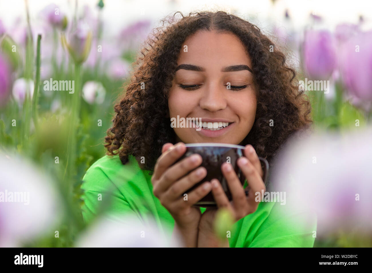 Razza mista bella African American biracial ragazza adolescente femmina giovane donna bere tazza di tè o caffè che indossa una maglietta verde nel campo della rosa Foto Stock