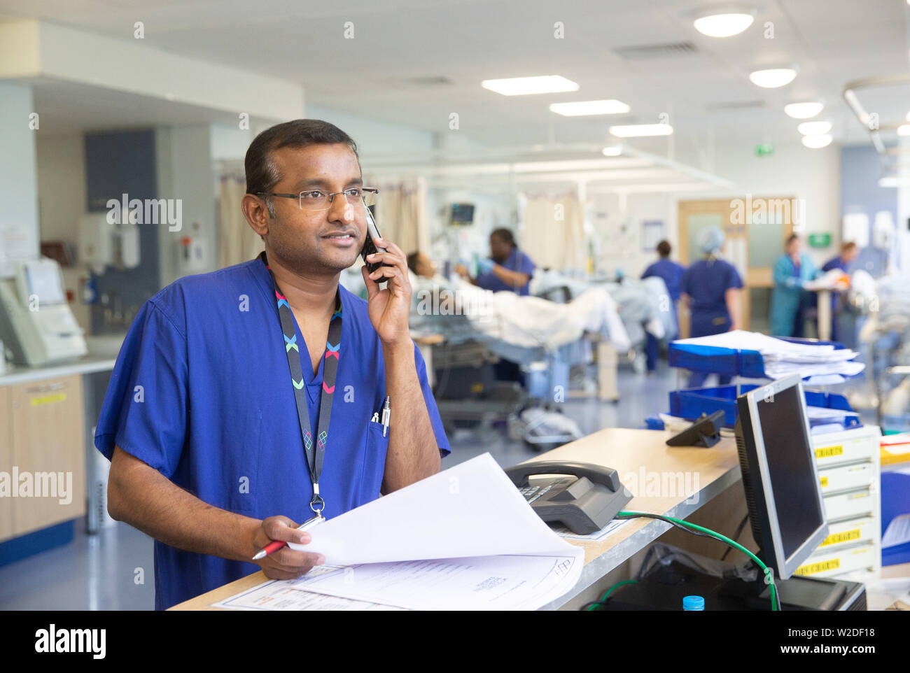 Maschio infermiera NHS in corrispondenza di una stazione di lavoro in un ospedale ha a che fare con le note paziente e informazioni per telefono. Foto Stock