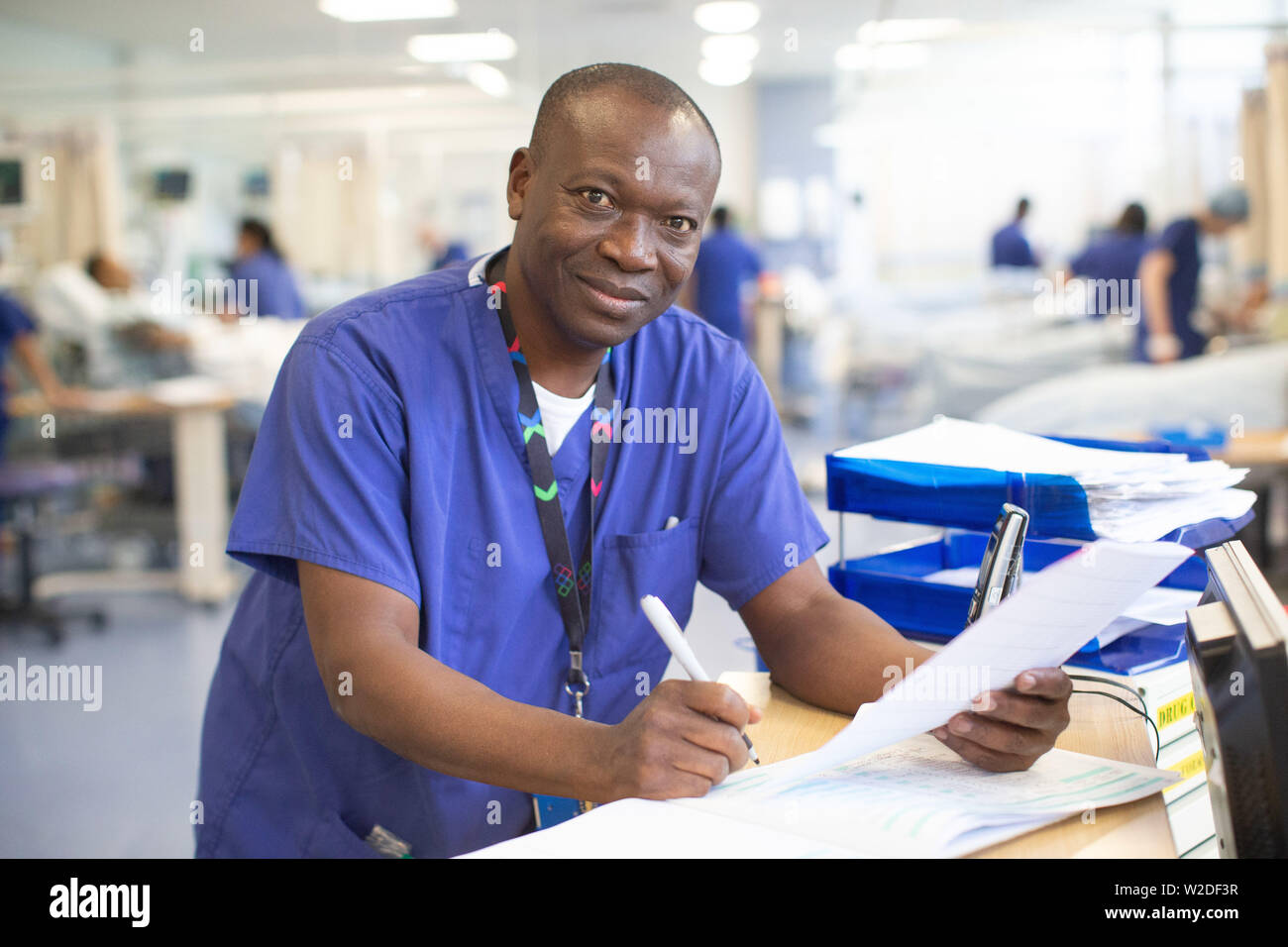 Maschio infermiera NHS in corrispondenza di una stazione di lavoro in un ospedale ha a che fare con le note paziente e informazioni per telefono. Foto Stock