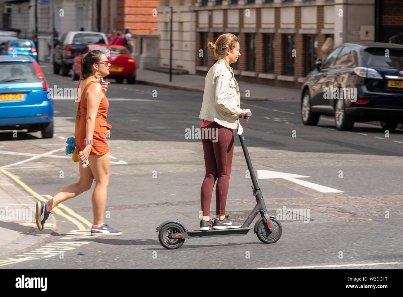 Femmina bianca caucasica che guida uno scooter elettrico fuori dal marciapiede utilizzando un passaggio pedonale di una strada a Londra, vicino a un pedone a piedi. Nessun cappello Foto Stock
