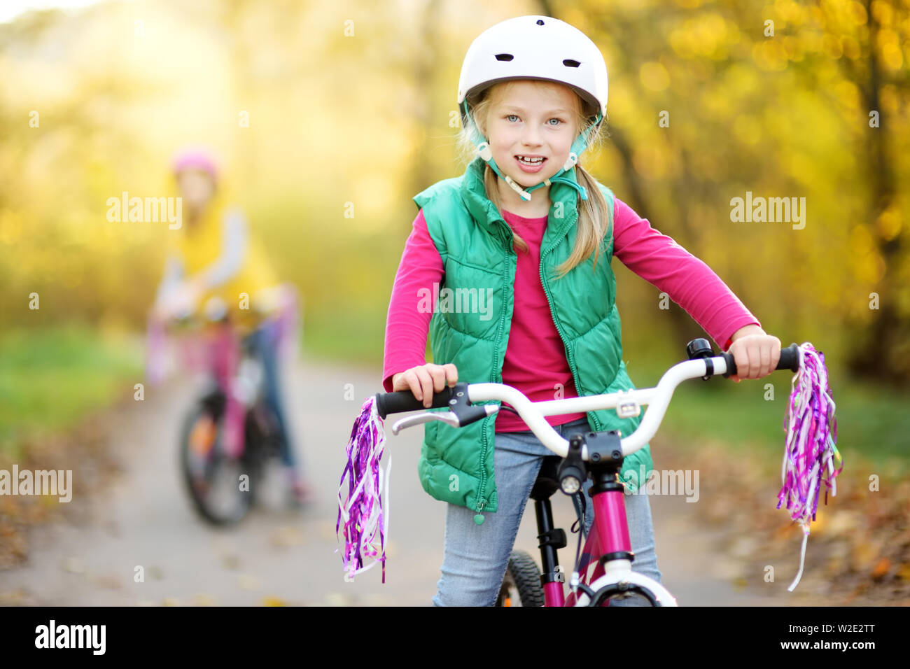 Carino piccole sorelle equitazione biciclette in un parco della città sulla soleggiata giornata autunnale. Attiva vacanza in famiglia con i bambini. Bambini indossare l'elmetto di sicurezza durante la guida di un bi Foto Stock
