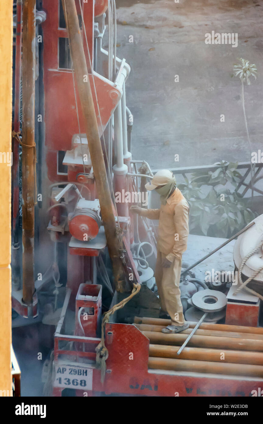 La polvere fine circonda il foro di trivellazione di un operatore addetto alla macchina che indossa una maschera facciale e il monitoraggio del rig, foratura per individuare acqua macinata, Hyderabad, India. Foto Stock
