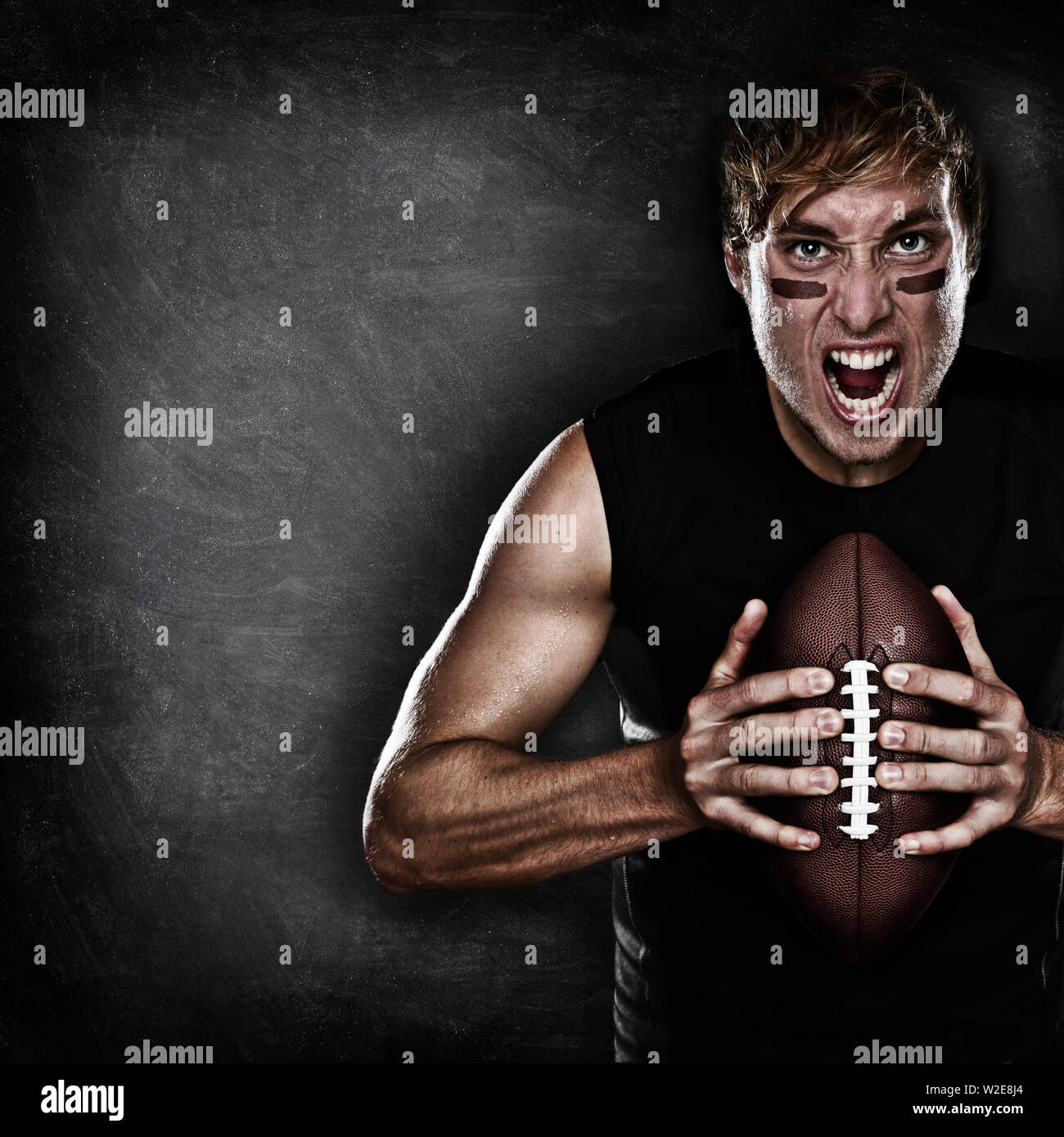 Giocatore di football ritratto aggressivo tenendo il football americano sulla lavagna nera sfondo con copia spazio per il testo o il design. Caucasica modello maschio nel suo 20s. Foto Stock