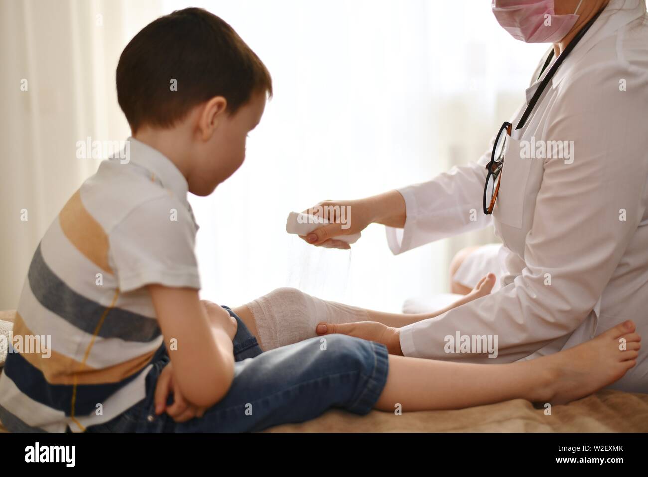 Un medico in una veste bianca bende il ginocchio di un bambino seduto di fronte a lei. Il ragazzo in attesa, piegato le sue braccia e orologi la scena. Foto Stock