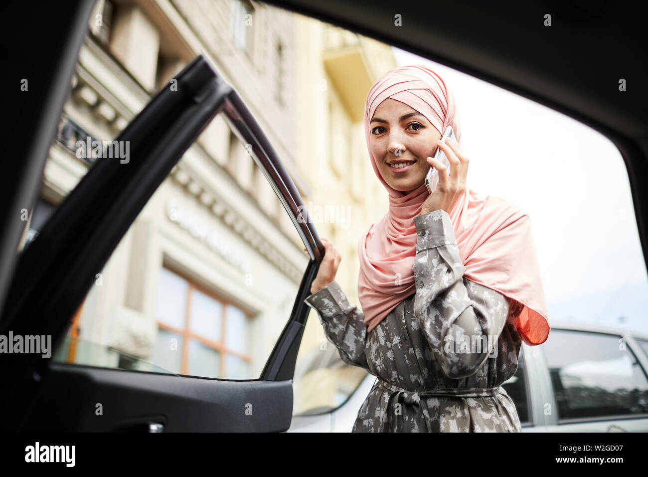 Allegro piuttosto giovane donna in hijab apertura porta auto e guardando camer Foto Stock