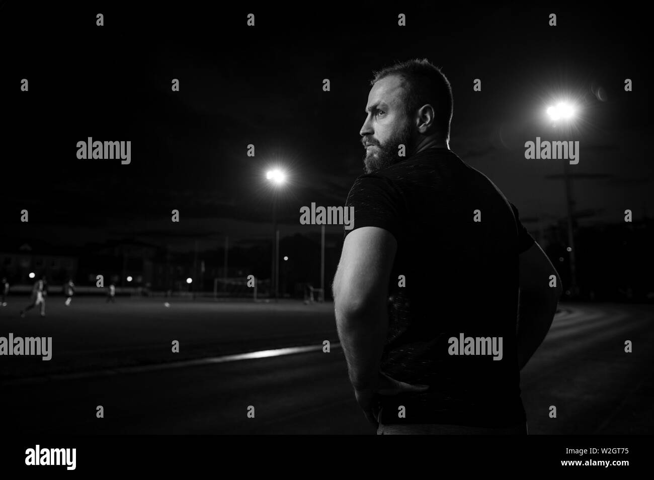 Persona tenace giovane uomo barbuto in t-shirt in piedi sul Crepuscolo stadio sportivo di guardare al gioco del calcio Foto Stock