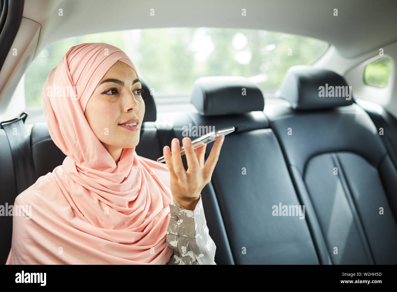 Contenuto fiducioso bella Arabian imprenditrice in rosa hijab seduto sul sedile posteriore di un taxi e la registrazione del messaggio vocale sul telefono Foto Stock