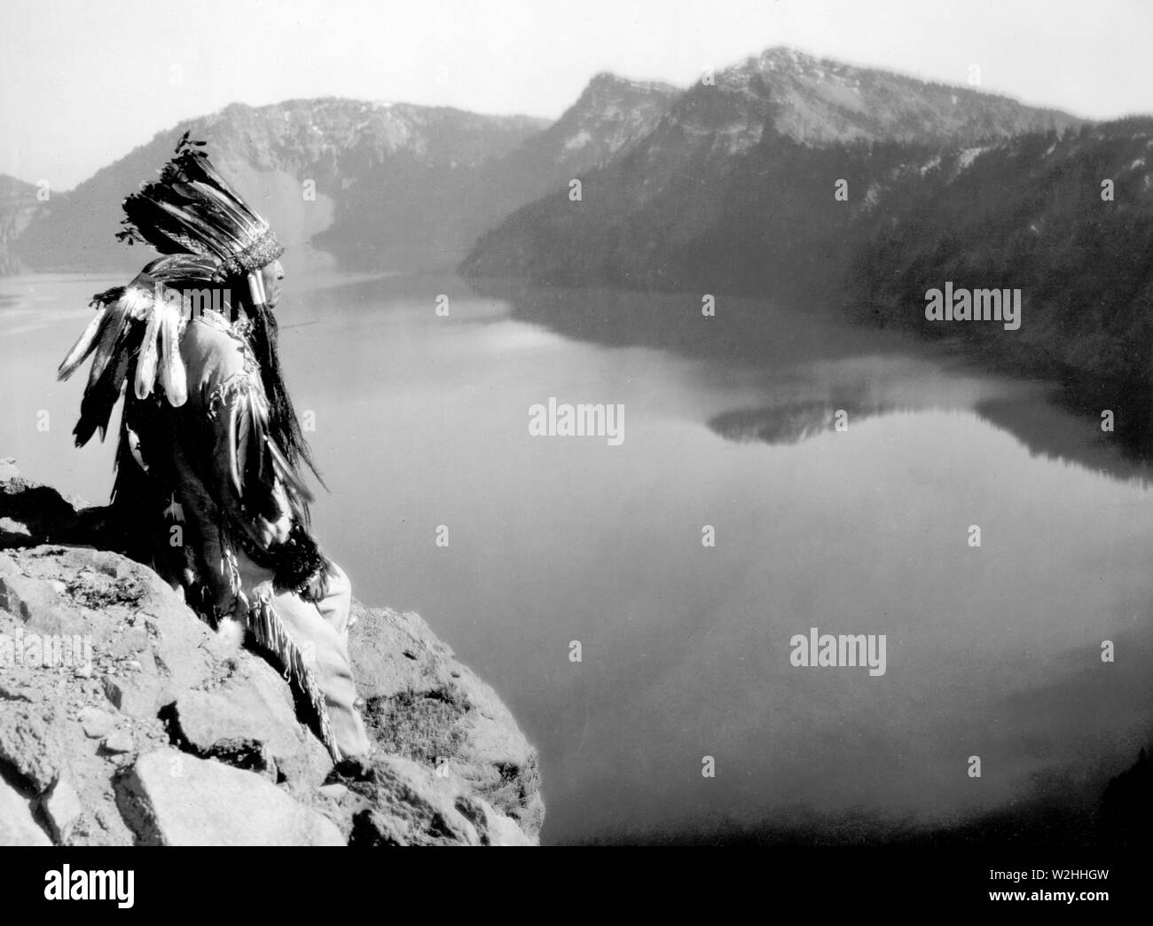 La fotografia mostra un American Indian uomo che indossa un headress, seduto su di una roccia overlocking di Crater Lake, Oregon Foto Stock