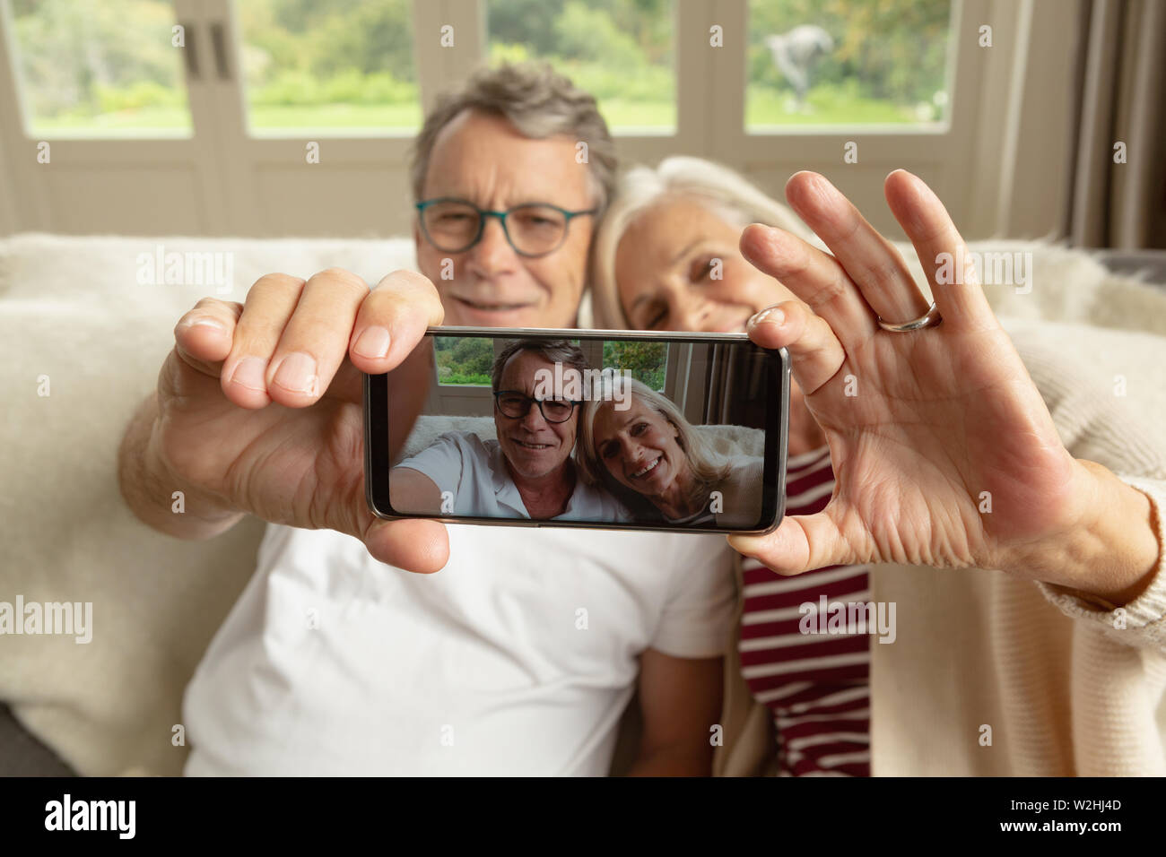 Active senior giovane seduto sul divano e prendendo selfie in una casa confortevole Foto Stock