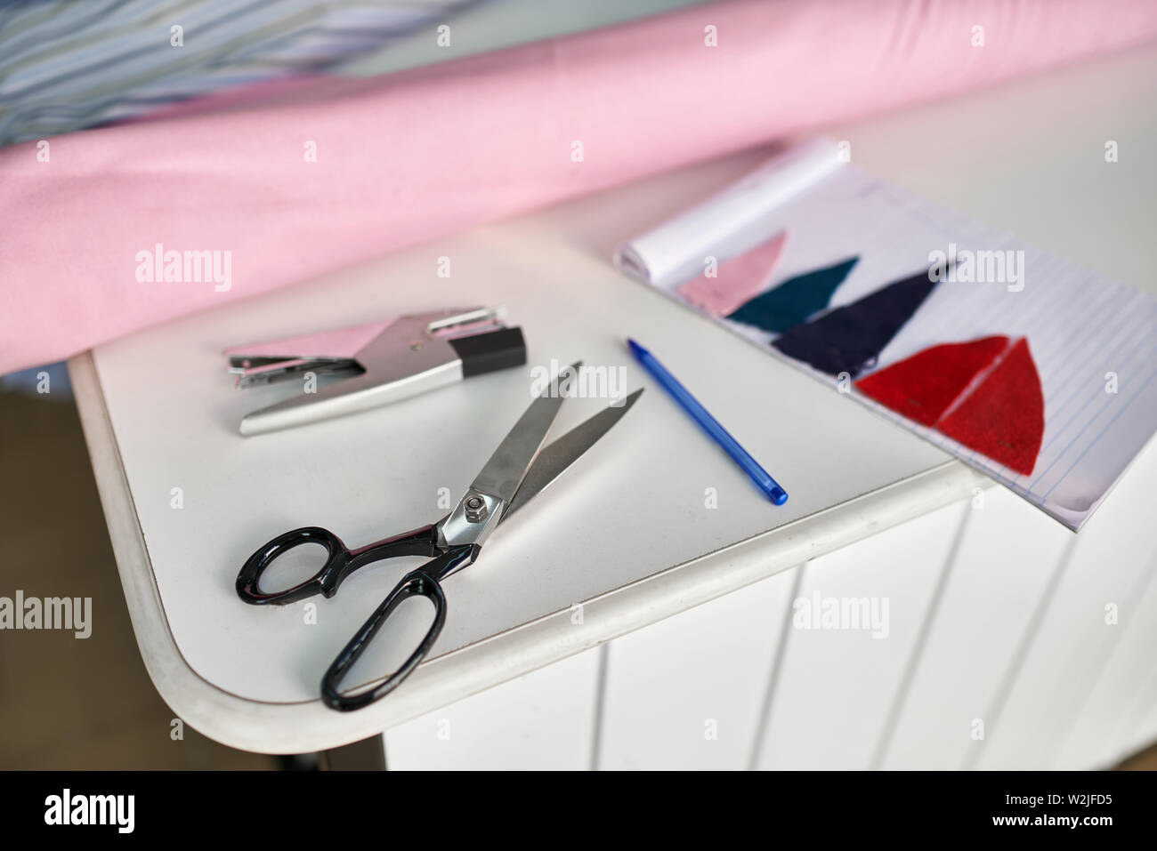 Forbici nero, cucitrice, penna blu, notebook con multicolore pezzi tessili sono sdraiato sul tavolo bianco all'interno. Non vi è una rosa rotolo di tessuto e una t Foto Stock