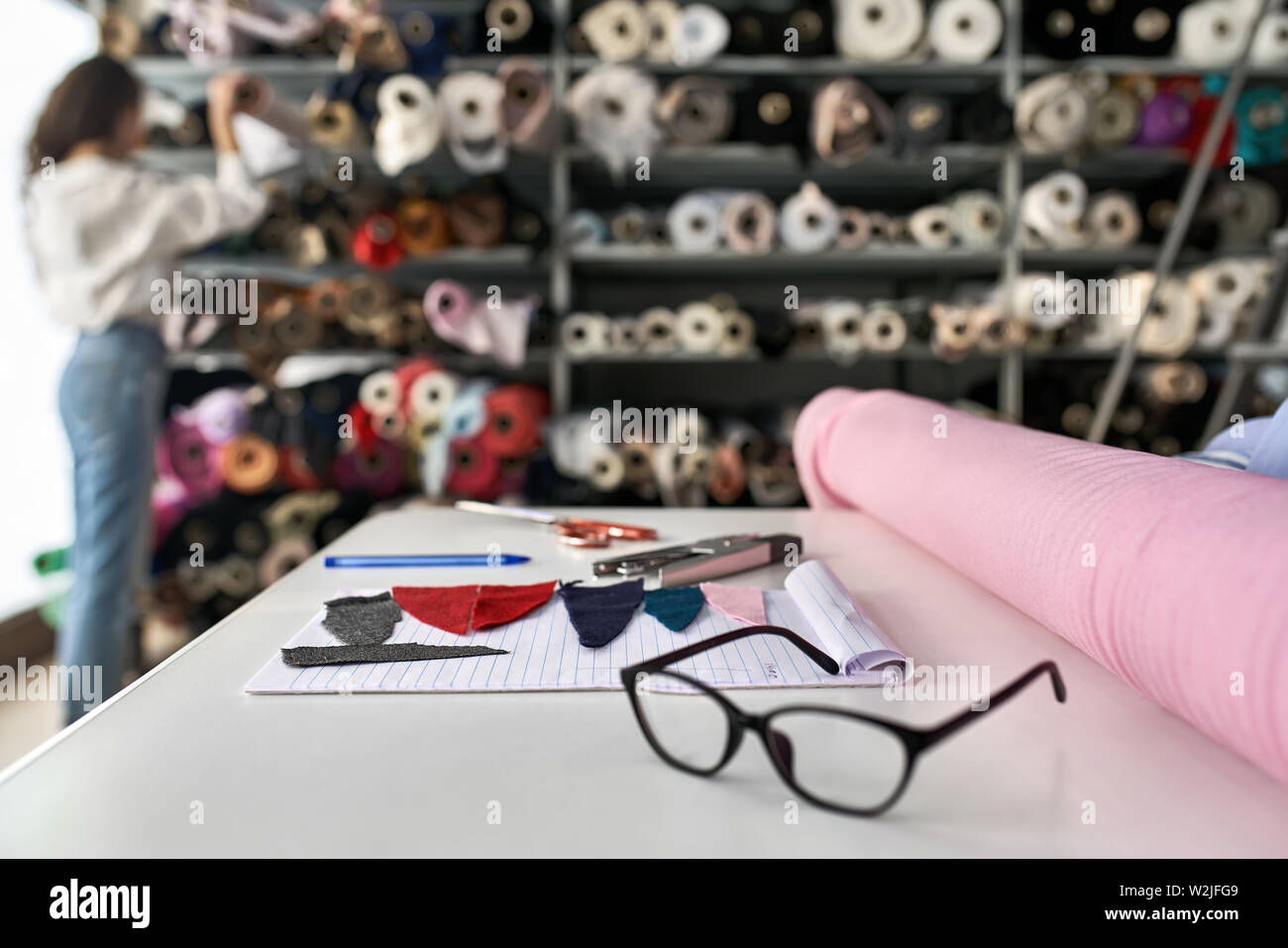 Ragazza sceglie un materiale da ripiani con multicolore di rotoli di tessuto all'interno di un magazzino. Di fronte a lei vi è tavolo bianco con pezzi di tessili e di un Foto Stock