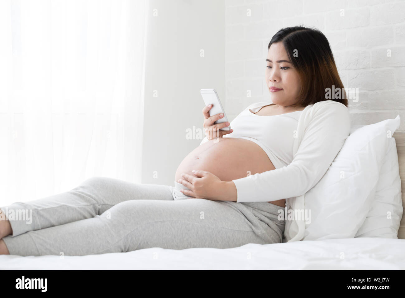 Asian donna incinta con lo smartphone e a mantenere aggiornate sul letto di casa utilizzando il telefono cellulare durante la gravidanza e i social media addiction concept Foto Stock