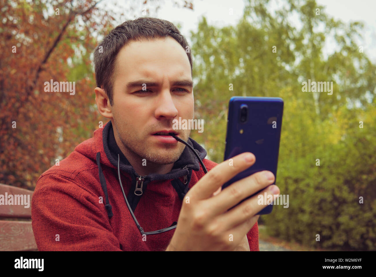 Uomo di pensiero curiosamente, tenendo in mano uno smartphone, seduti all'aperto in giardino. Un giovane ragazzo indignantly guarda lo schermo del suo smartphone. La lettura di bad Foto Stock
