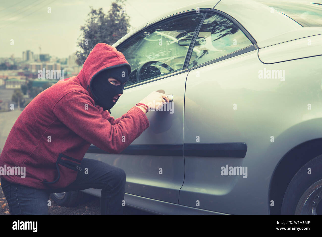 Uomo Mascherato in abbigliamento casual furtivamente si preleva la serratura di un veicolo su una città sullo sfondo. Ladro maschio furtivamente picklocks la portiera del veicolo. Programma di intercettazione con cautela Foto Stock