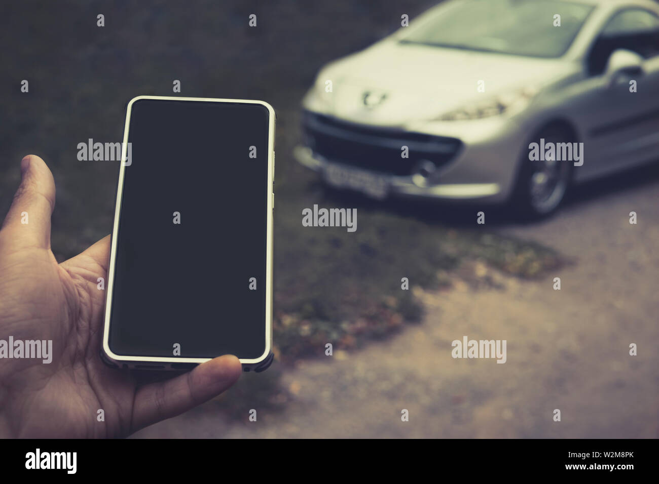 Smartphone con schermo nero nel maschio palm a veicolo parcheggiato sfondo. Avviamento del motore del servizio mobile. Telefono cellulare invia segnali per sbloccare il veicolo Foto Stock
