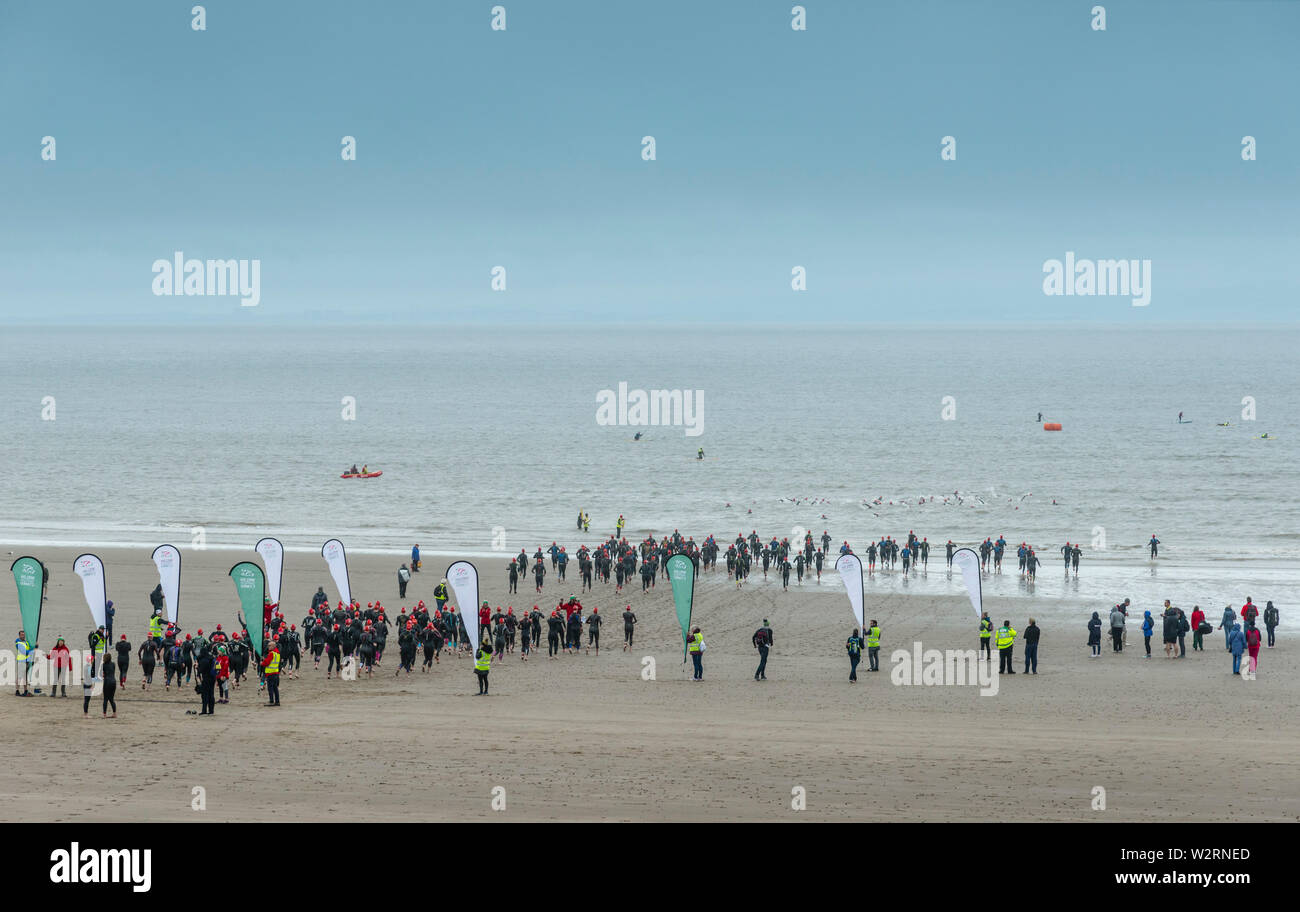 Sotto le nuvole scure concorrenti nel 2019 Barry Island sprint triathlon corrono attraverso la spiaggia di sabbia e nel mare all'inizio della gara. Foto Stock