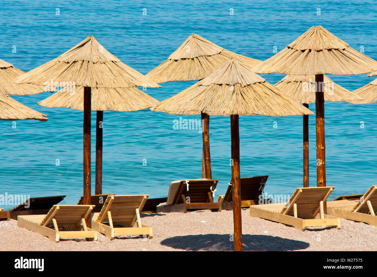 Ombrelloni di paglia e lettini in legno su una spiaggia deserta e acqua turchese in background Foto Stock