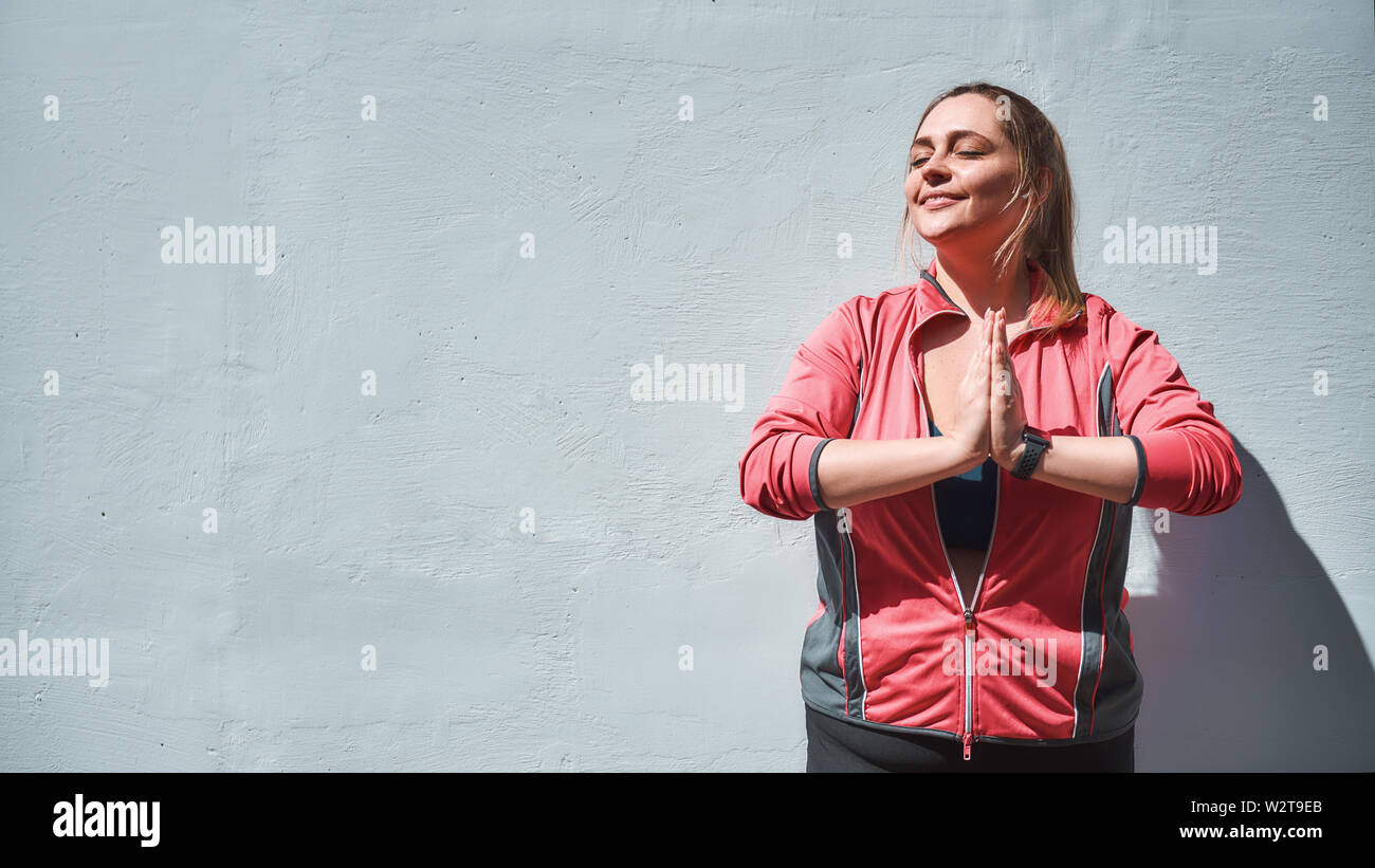 Equilibrio di vita. Donna calma e bella più taglia in abiti sportivi facendo yoga mentre si sta contro parete con occhi chiusi. Concetto di yoga. Perdita di peso. Donne grumi Foto Stock