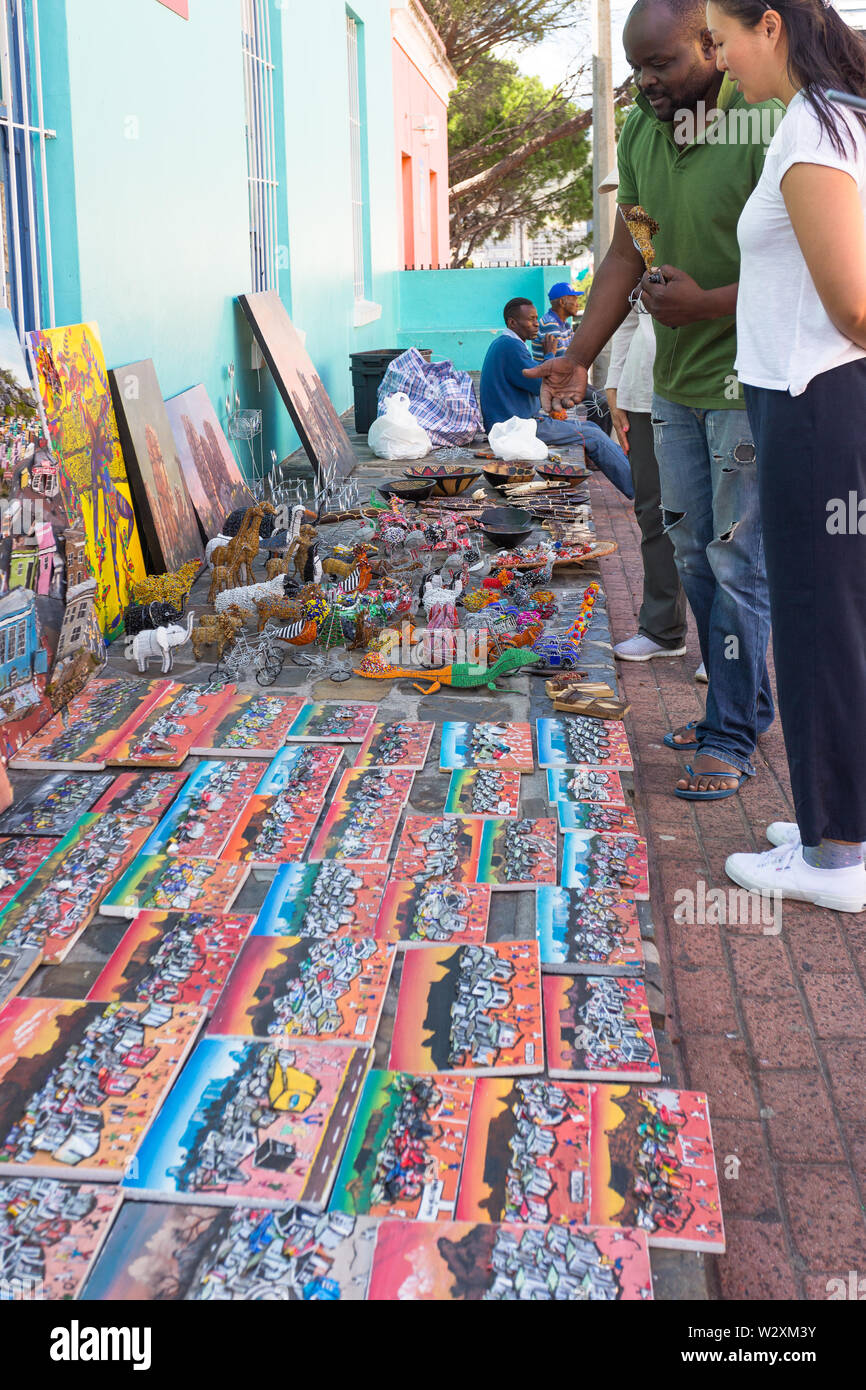 Donna asiatica navigazione turistica tradizionale africana di curiosità o souvenirs acquistare da un Africano nero venditore ambulante in Bo Kaap, Cape Town, Sud Africa Foto Stock