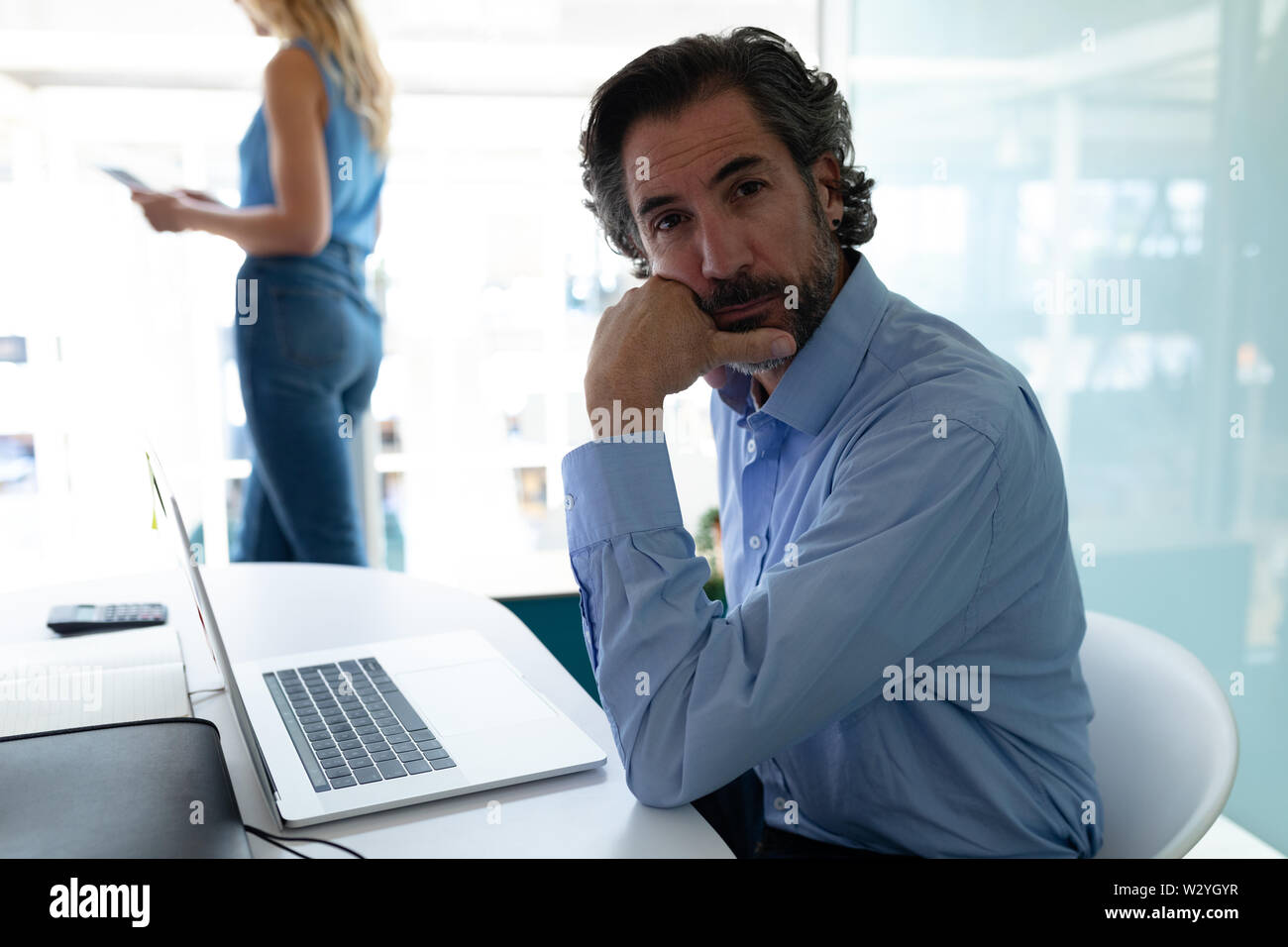 Imprenditore con mano sul mento guardando la fotocamera mentre imprenditrice in piedi in background in ufficio Foto Stock