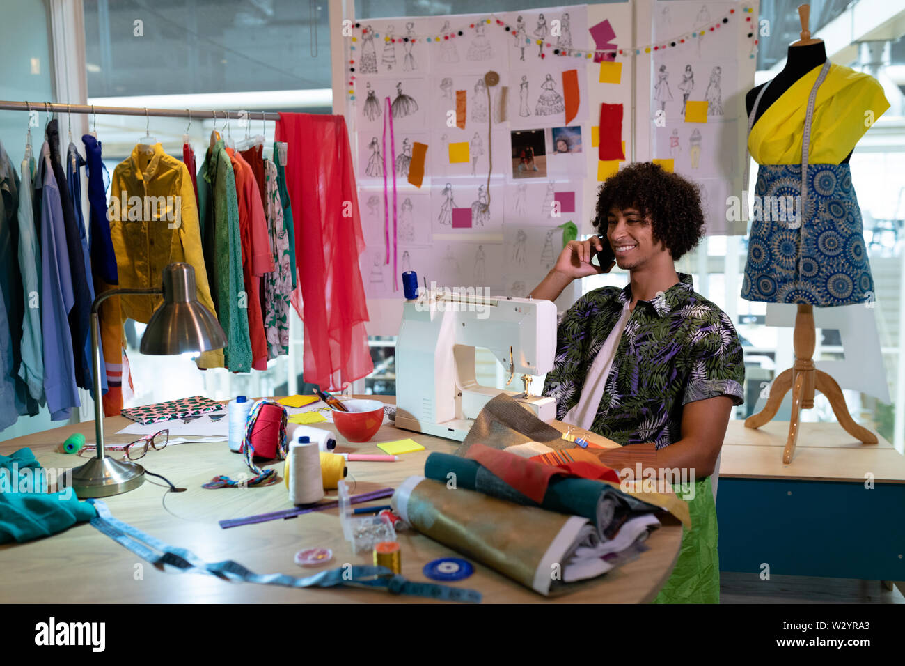 Maschio di designer di moda parlando al telefono cellulare mentre si utilizza la macchina per cucire Foto Stock
