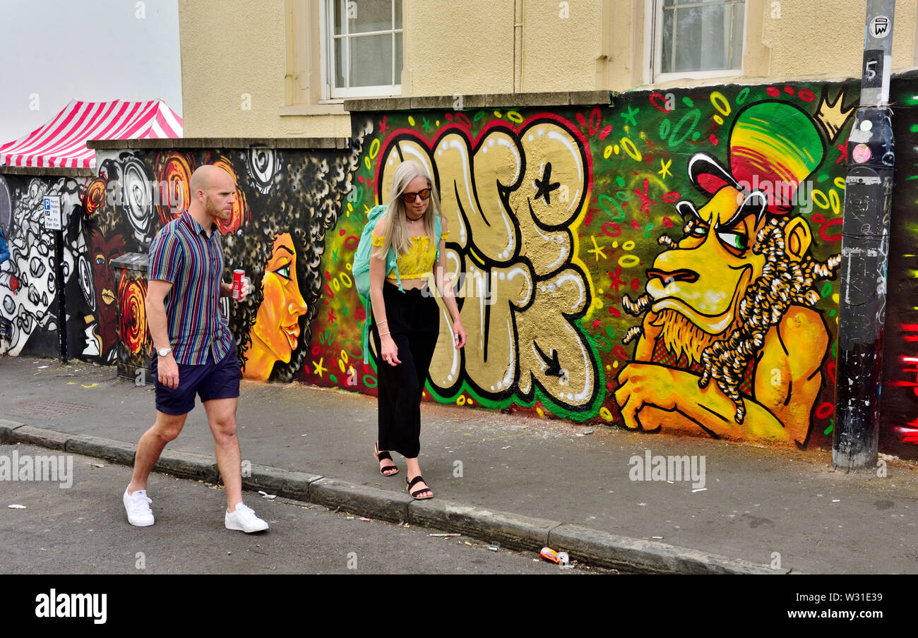 Due persone a piedi lungo la strada di San Paolo Bristol con arte di strada dietro durante il tempo di carnevale Foto Stock