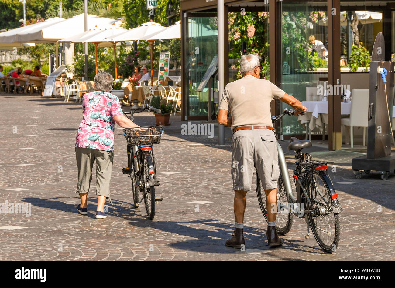 Il LAGO DI GARDA, Italia - Settembre 2018: uomo maturo e la donna wheeling biciclette passato ristoranti sul lago di Garda sul Lago di Garda. Foto Stock