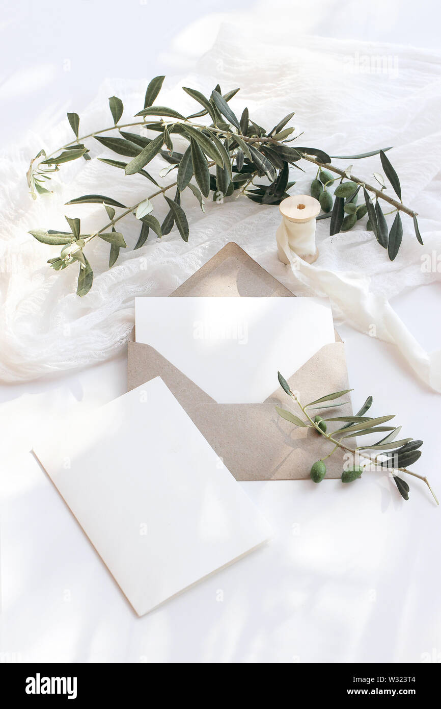 Estate wedding stationery mock-up scena. Biglietti vuoti, busta, di rami di olivo e nastro di seta. Sfondo bianco con cotone tabella runner in Foto Stock