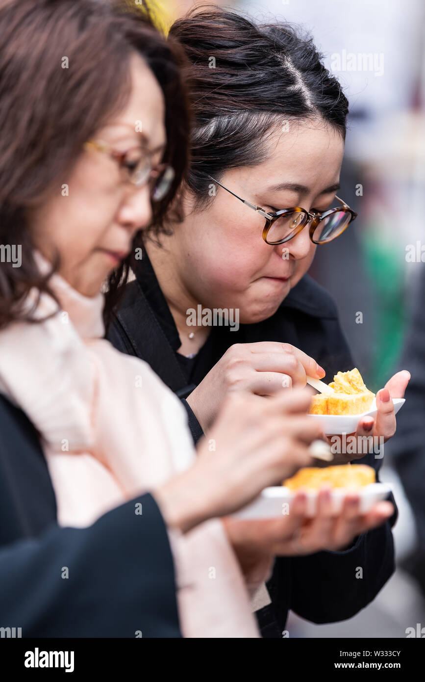 Tokyo, Giappone - 30 Marzo 2019: vista laterale di due Asian Le donne cinesi mangiare giapponese tamagoyaki uova frittata arrotolata in del pesce di Tsukiji street mercato esterno Foto Stock
