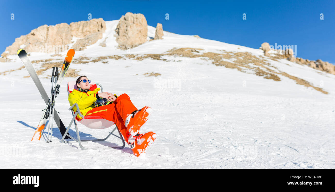 Immagine di sport uomo seduto sulla sedia accanto a sci e bastoni sullo sfondo di montagne innevate Foto Stock