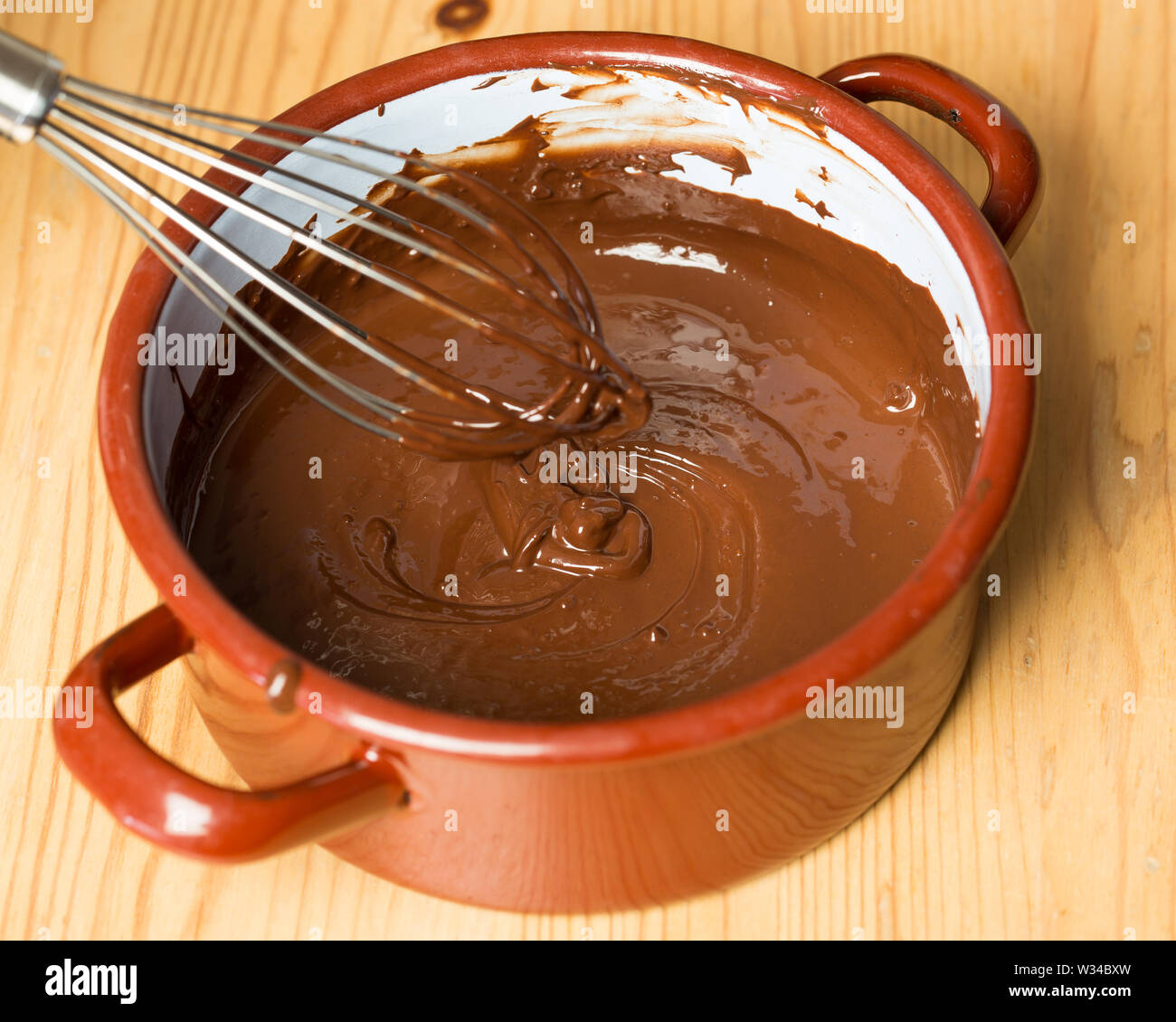Pentola con frusta e cioccolato fuso su pannello di legno Foto Stock