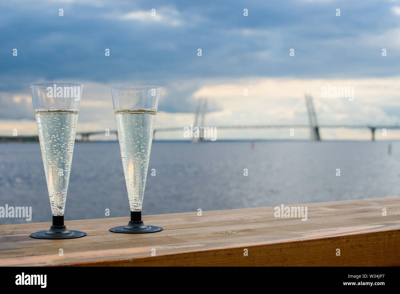 Due bicchieri di plastica di spumante su una superficie in legno sullo sfondo della città, il grande ponte, il mare e il cielo scuro. Foto Stock