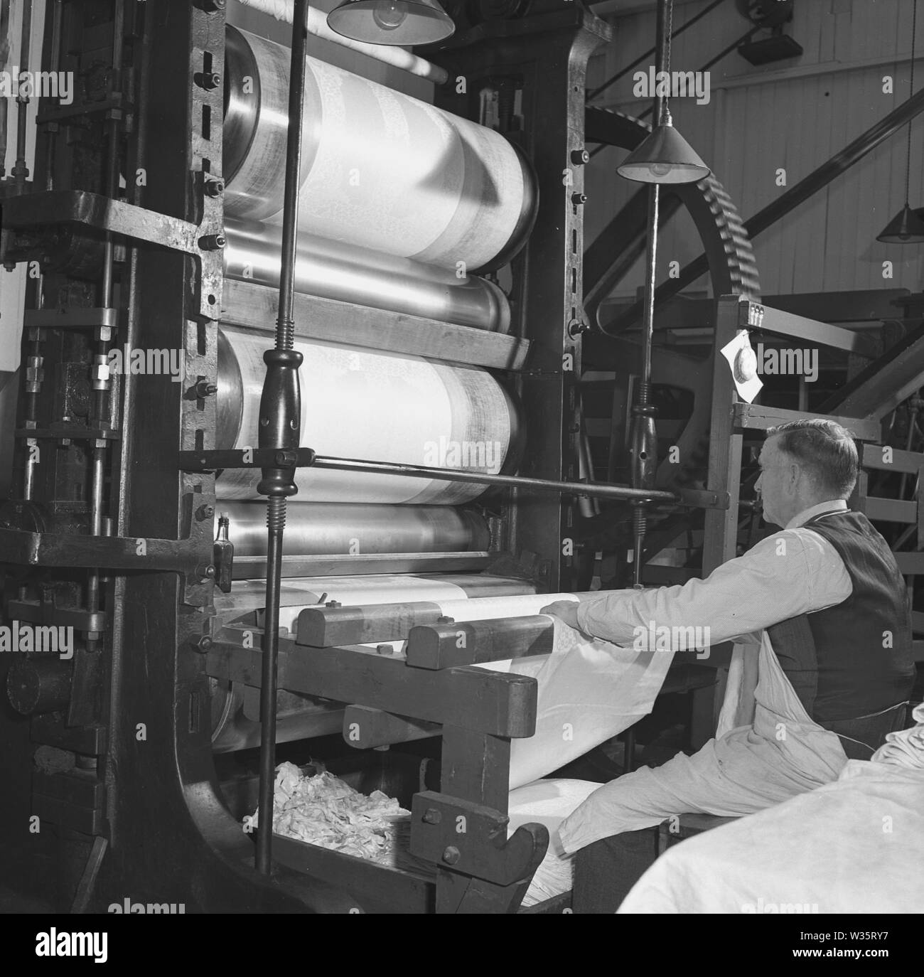 Anni '1950, un lavoratore di sesso maschile anziano che sovrintende a fogli di lino di nuova produzione in una grande macchina per la stampa a rulli in una fabbrica tessile, l'Irlanda del Nord, Foto Stock