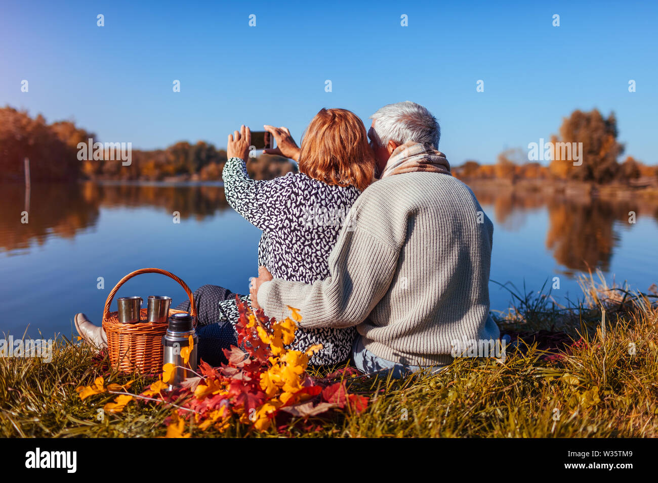 Coppia senior tenendo selfie sul telefono mentre avente picnic sulle rive del lago d'autunno. Felice l'uomo e la donna per godersi la natura e avvolgente Foto Stock