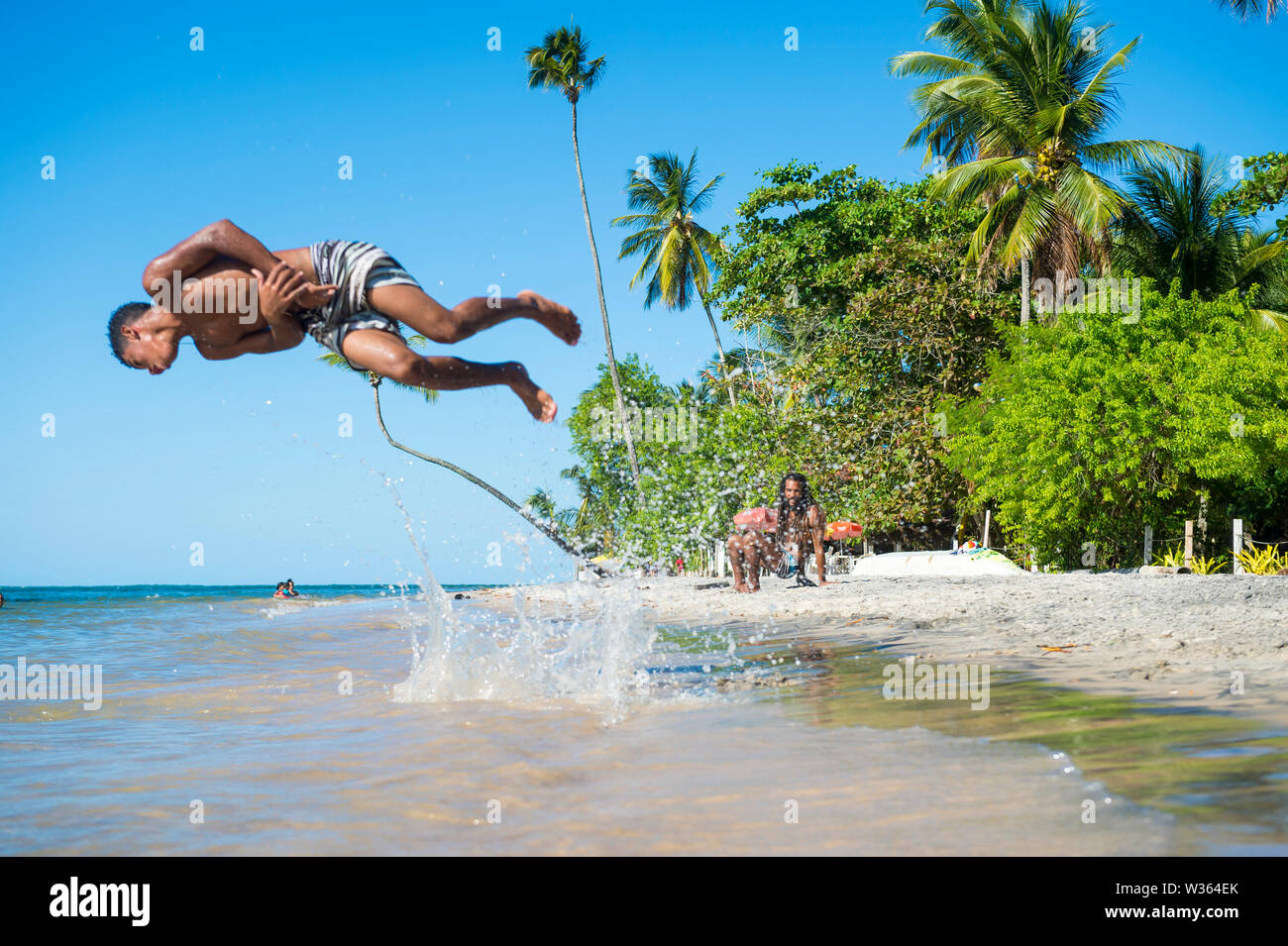 CAIRU, Brasile - Marzo 2018: un giovane brasiliano uomo fa un acrobatico flip in mare contro uno sfondo di ondeggianti palme tropicali. Foto Stock