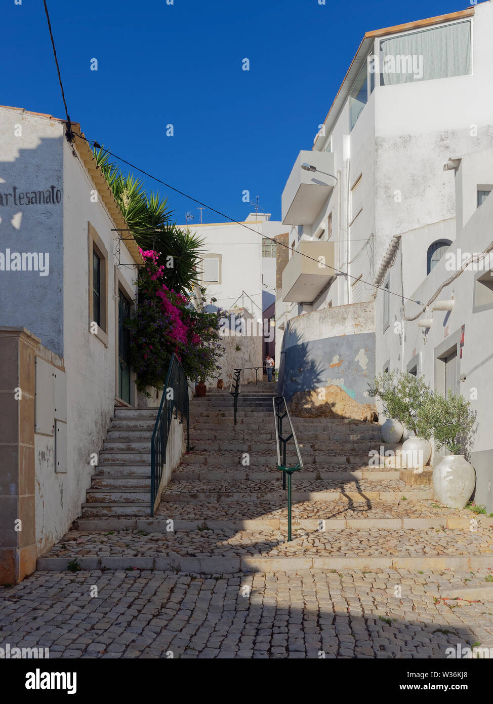 La ricerca di una vecchia strada di ciottoli con gradini in pietra e parapetti con una persona che cammina verso il basso tra le case a schiera. Estoi, Portogallo. Foto Stock
