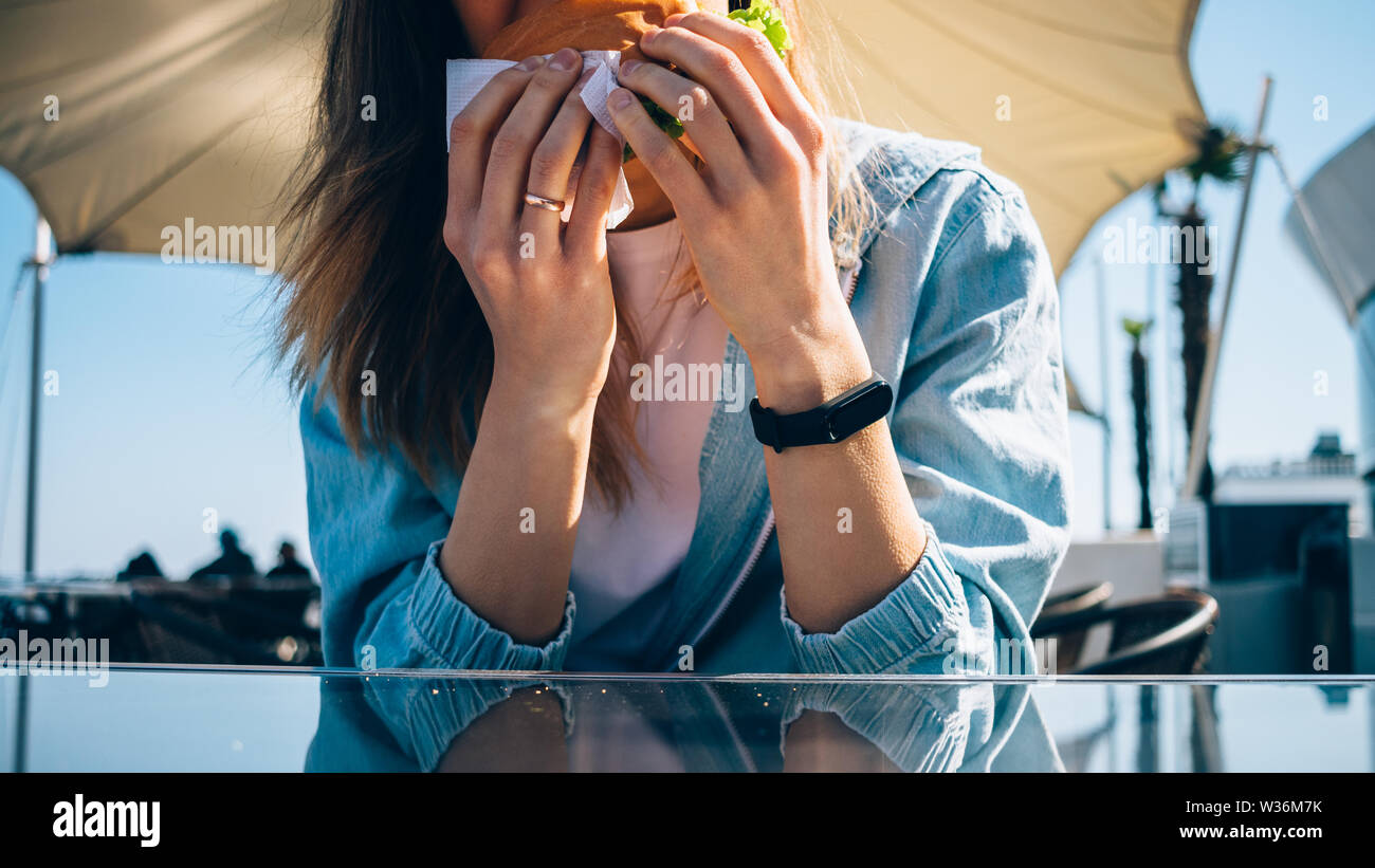 Close-up di giovane donna mangiare hamburger in seduta outdoor cafe a giornata di sole. Foto Stock